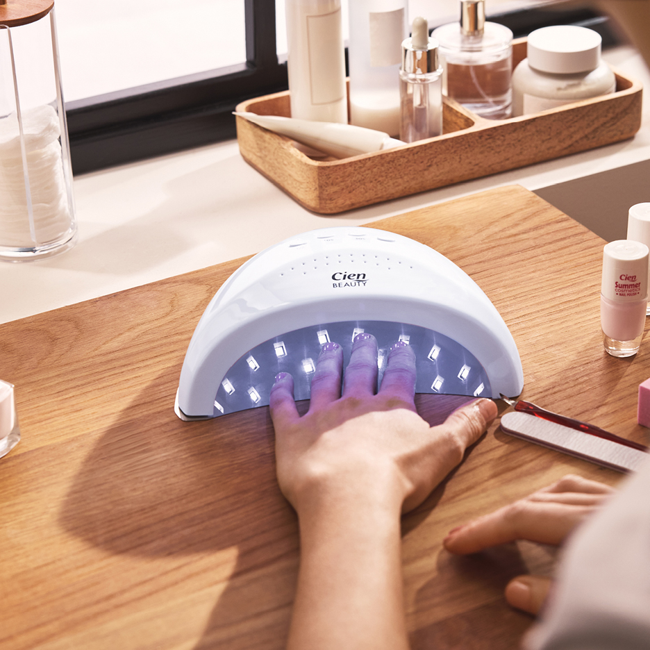 En hand under en vit UV-lampa för naglar, med nagellack och hudvårdsprodukter i bakgrunden.