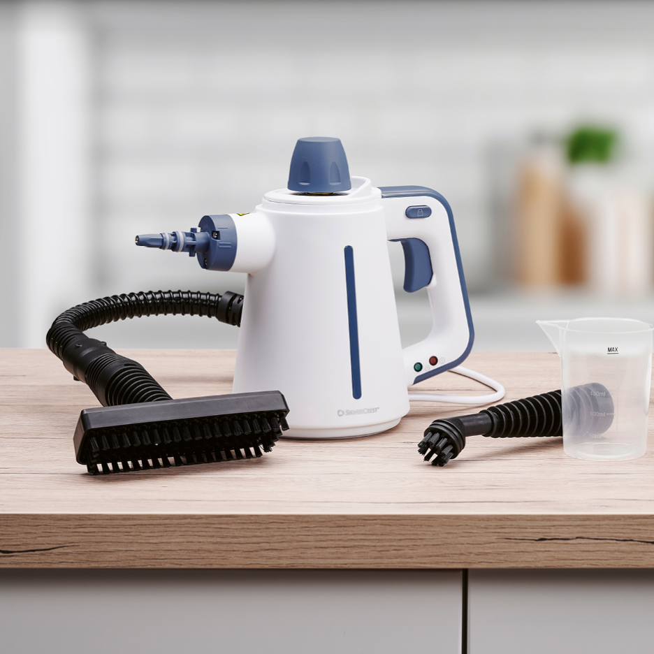 Handhållen ångtvätt med tillbehör som borstar och mätkopp på en träbänk.