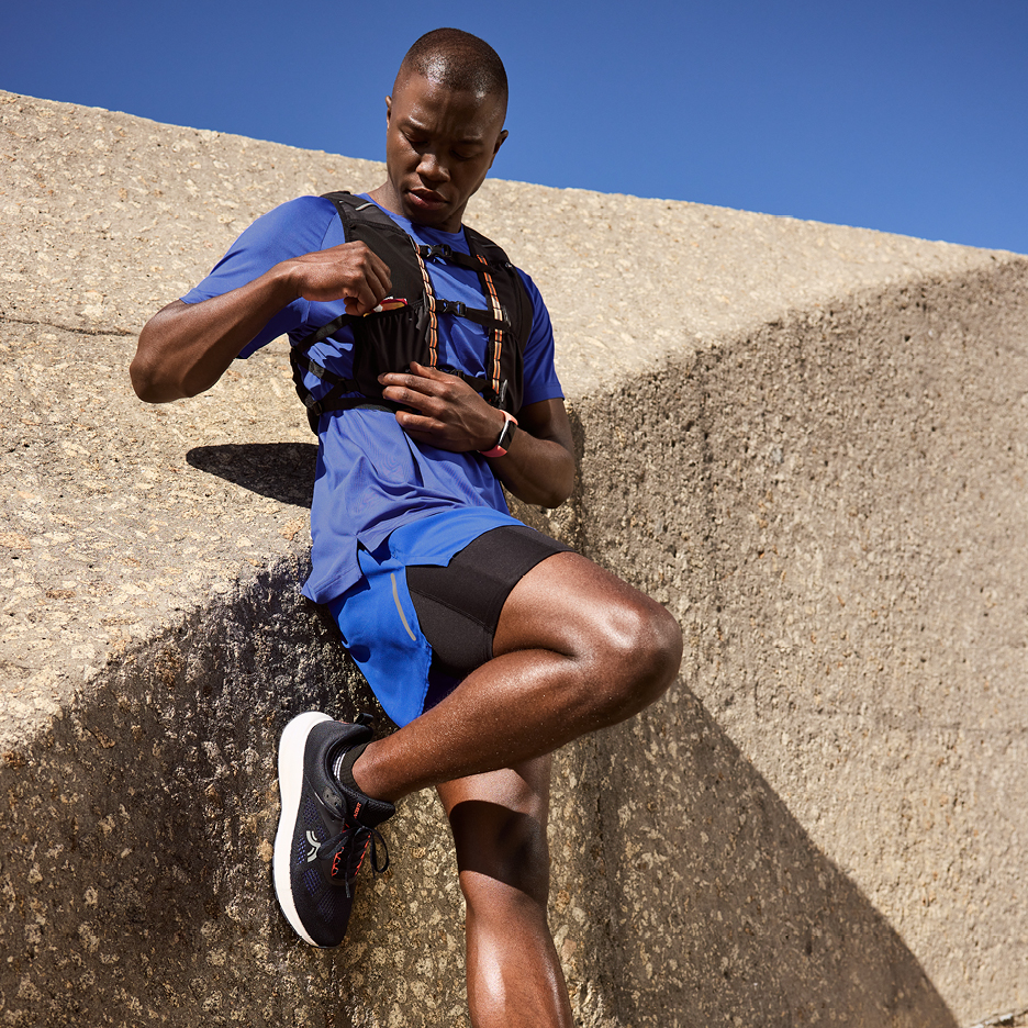 Man i blå löparutrustning med löparväst och löparskor, lutar sig mot en betongvägg.