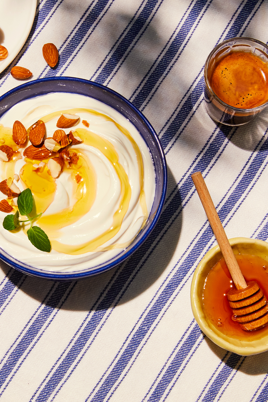 Yoghurt med honung och mandel, en kopp espresso och en skål med honung på en randig duk.